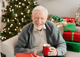 Elderly man sitting beside a Christmas tree drinking a cup of coffee with a nicely wrapped Christmas present on his lap.