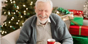 Elderly man sitting beside a Christmas tree drinking a cup of coffee with a nicely wrapped Christmas present on his lap.