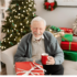 Elderly man sitting beside a Christmas tree drinking a cup of coffee with a nicely wrapped Christmas present on his lap.