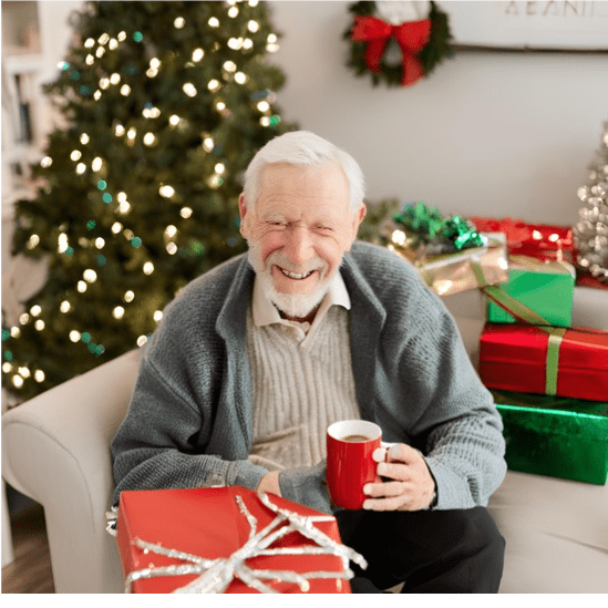 Elderly man sitting beside a Christmas tree drinking a cup of coffee with a nicely wrapped Christmas present on his lap.